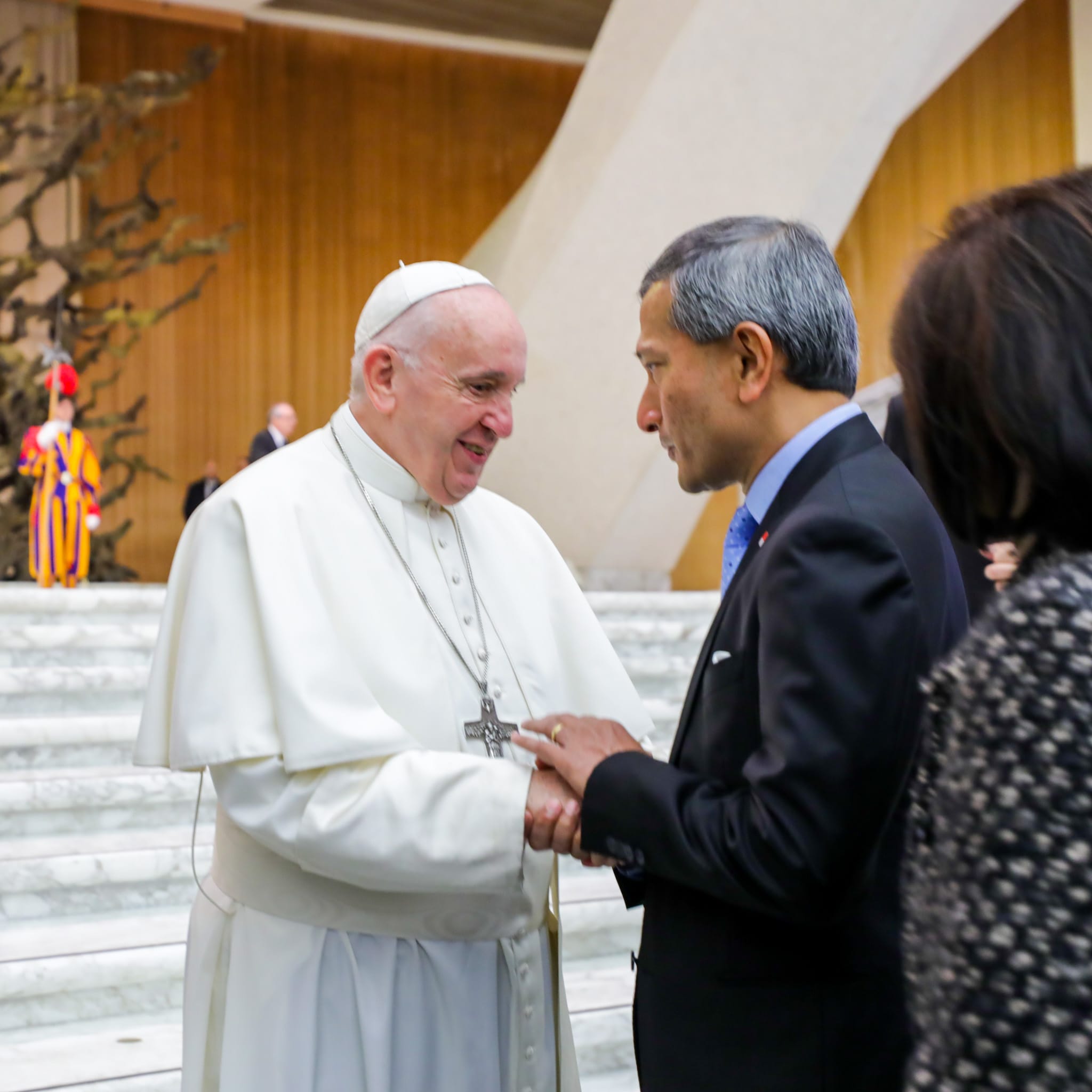 Pope Francis in white garb shaking hands with a man in a suit in a large hall.